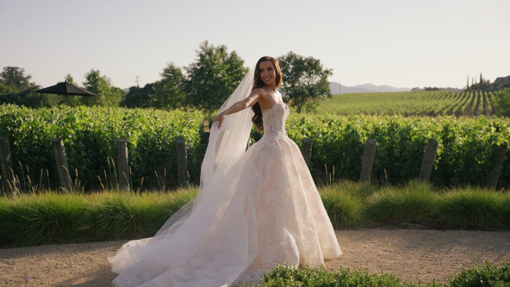 Full-length profile photo of a smiling bride twirling on a dirt path next to lush vineyard rows at Sunstone Winery, holding out her long tulle veil and A-line wedding dress.
