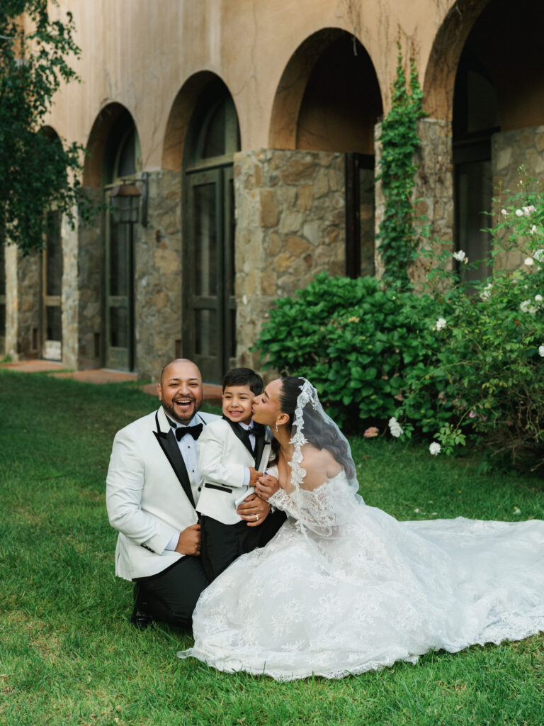 Bride in a lace gown and groom in a white tuxedo jacket kneeling on a green lawn with a young boy against a rustic stone estate villa with arches.
