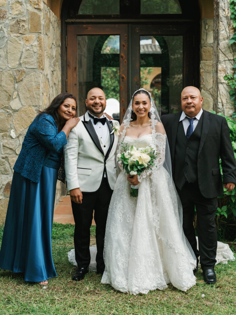 Elegant family wedding portrait at a European-style stone villa, featuring a bride in a lace off-the-shoulder gown and a groom in a sharp white tuxedo jacket with parents.