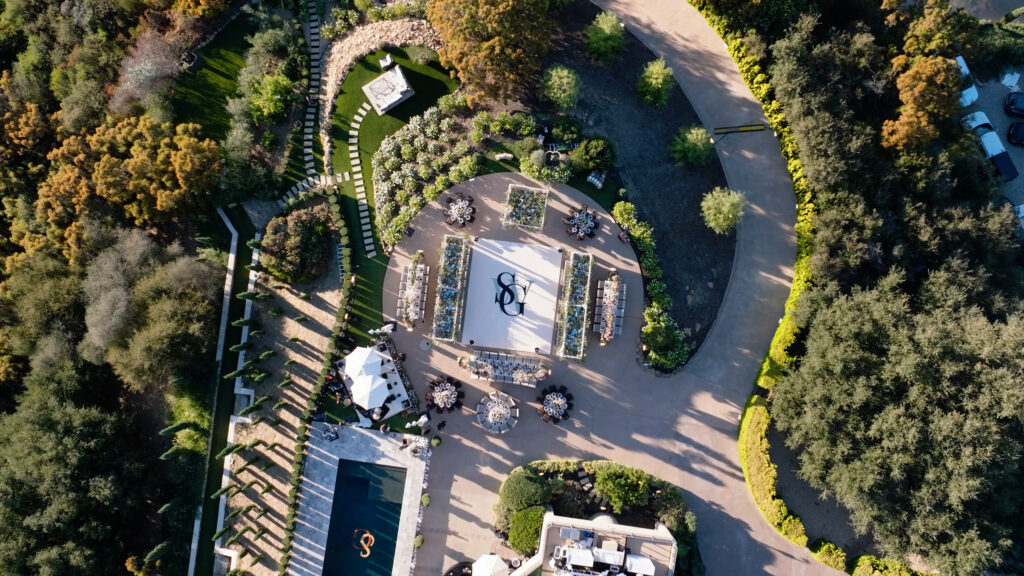 Birds-eye drone photography of a luxury wedding reception layout at the Pool Rest Area of Greystone Mansion, featuring a white custom dance floor with a monogram logo, banquet tables, and guest seating surrounded by lush estate gardens and stone walkways.
