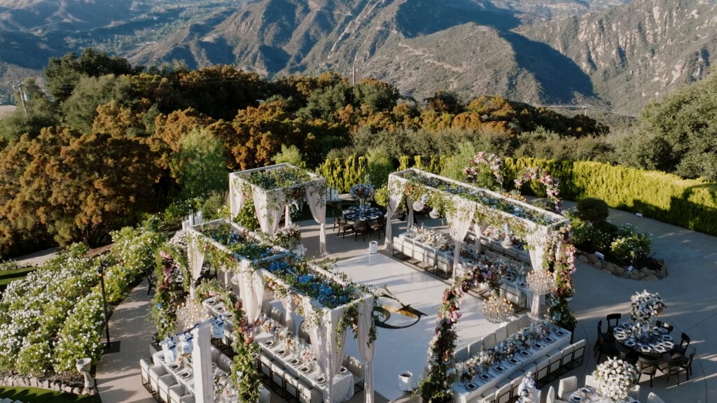 High-angle shot of an opulent wedding reception layout at the Formal Gardens of Greystone Mansion, featuring custom floral-covered structures, a white dance floor with a custom monogram, and a majestic mountain backdrop.