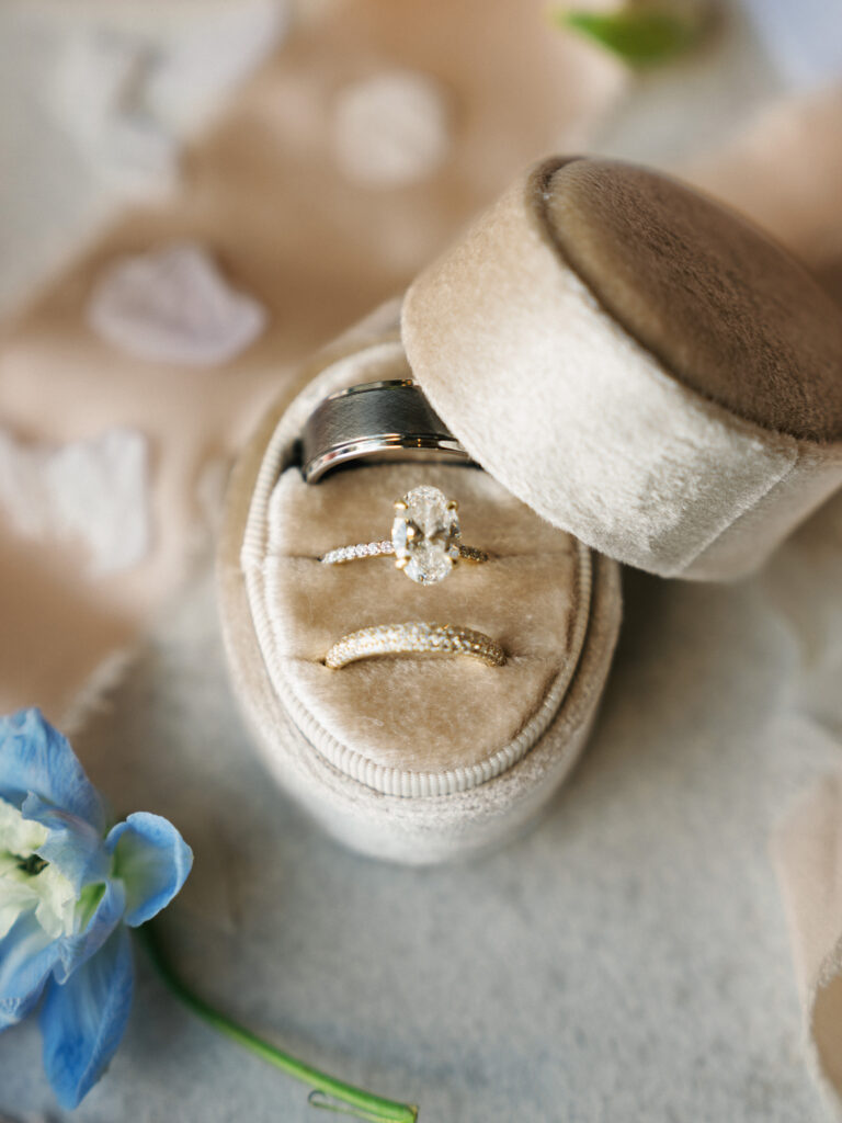 Oval cut diamond engagement ring with a pavé band and gold rings inside a beige velvet box, creating soft wedding detail photos with a blue flower.