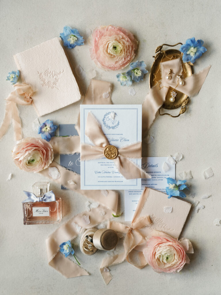 Pastel fine art wedding invitation suite flat lay featuring peach ranunculus, blue delphinium, silk ribbons, and Miss Dior perfume for romantic wedding detail photos.