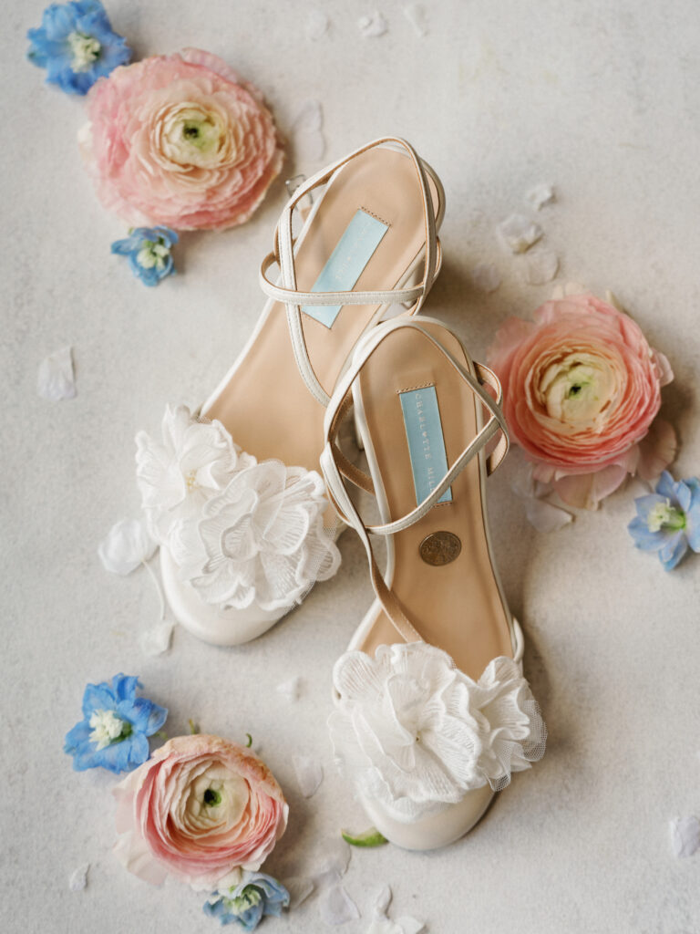 White tulle flower Bella Belle bridal shoes styled on a textured neutral surface with peach and blue blossoms for high-end wedding detail photos.