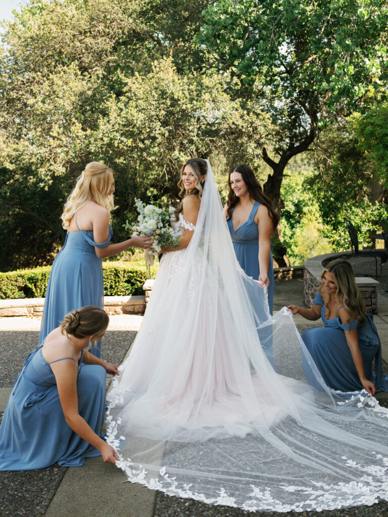 Bridesmaids in dusty blue dresses fluffing a long lace-trimmed cathedral veil for a bride in an off-the-shoulder A-line wedding gown outdoors.