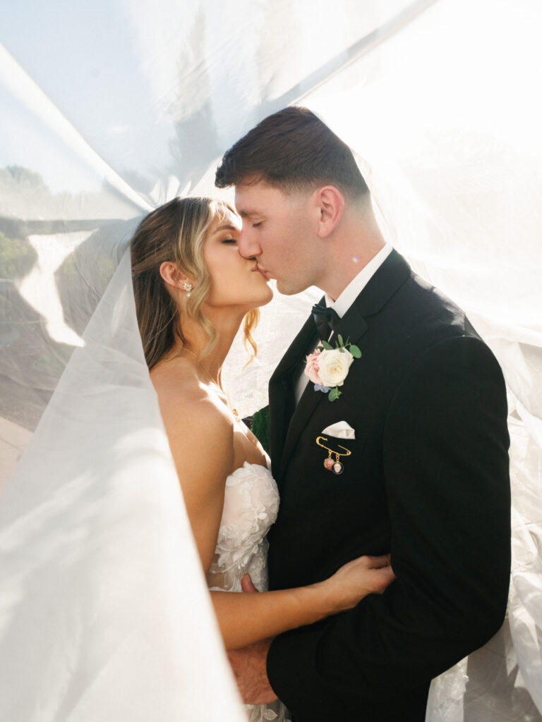 Romantic newlyweds kissing under a sheer white wedding veil, featuring a groom in a black tuxedo with a pastel floral boutonniere.