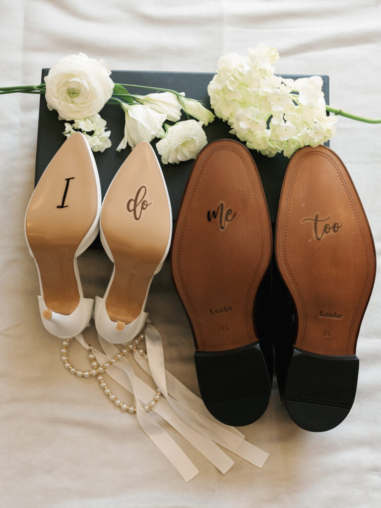 White bridal heels and black groom dress shoes showing custom I Do Me Too sole calligraphy next to white ranunculus flowers.