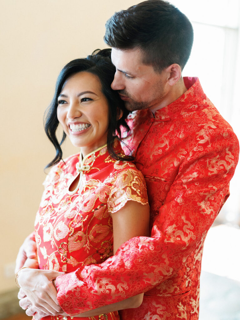 Candid portrait of a bride in a traditional red Chinese Cheongsam hugging her groom at a multicultural wedding.