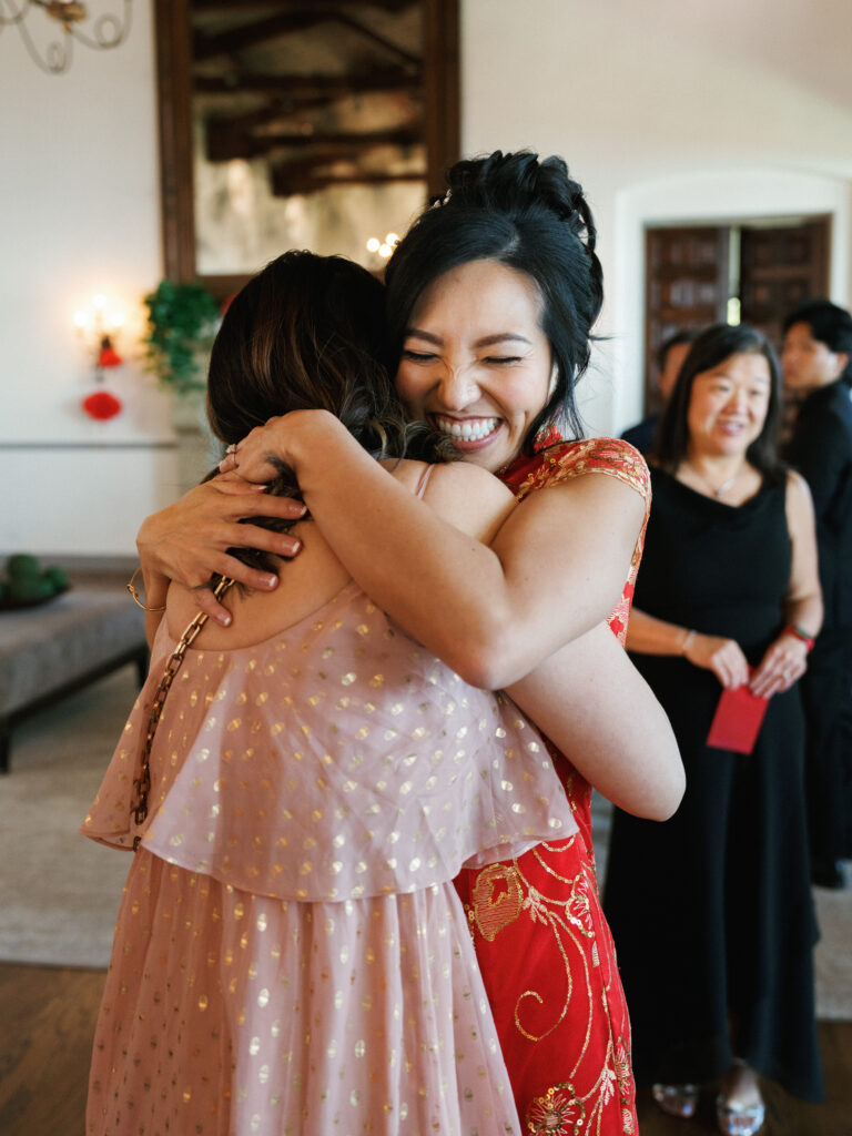 A bride in traditional red Chinese wedding attire embracing a guest during a multicultural wedding celebration.