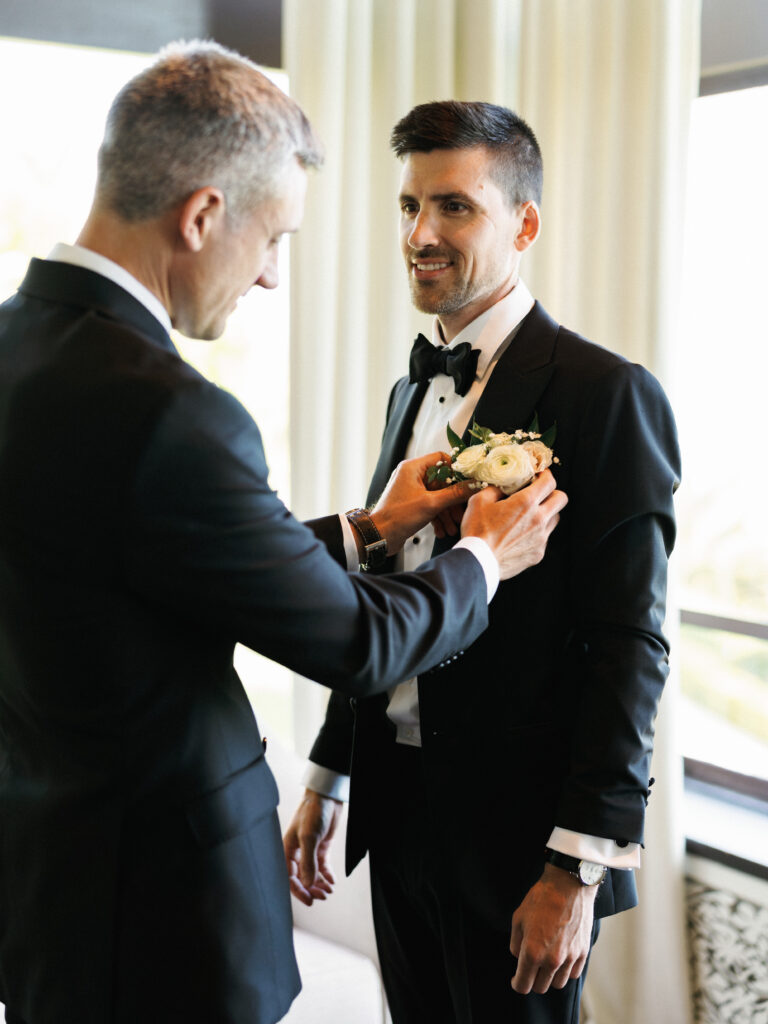 Best man adjusting a white rose boutonniere on the groom's black tuxedo lapel during wedding preparation.
