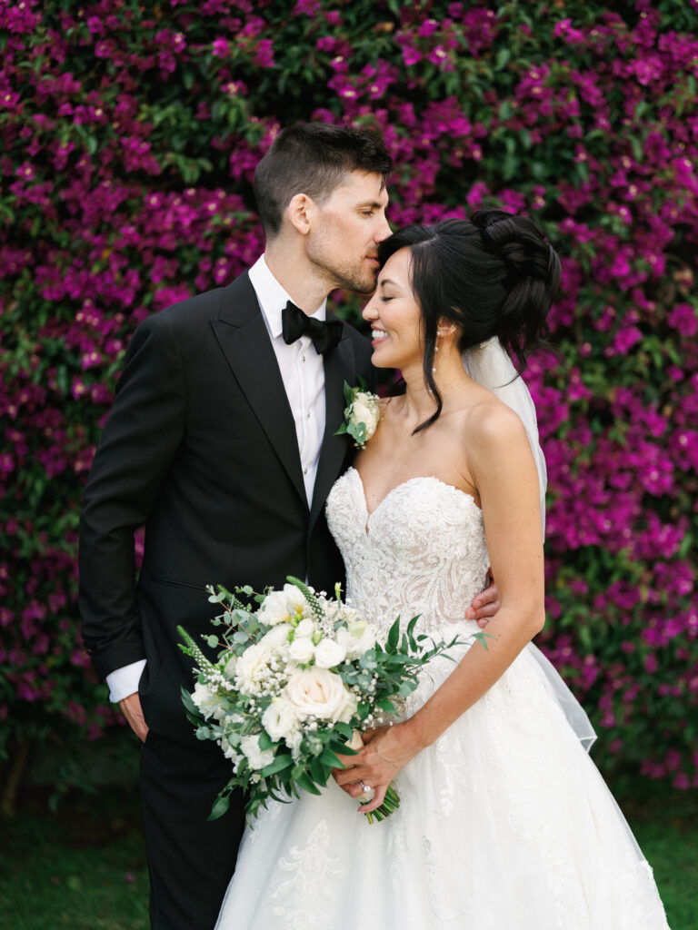 A groom kisses his bride’s forehead in front of a vibrant purple bougainvillea wall at a luxury Santa Barbara estate wedding.
