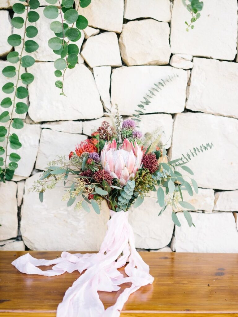 Boho luxury wedding bouquet featuring a large pink king protea, eucalyptus leaves, and long blush silk ribbons resting on a wooden bench against a rustic white stone wall.