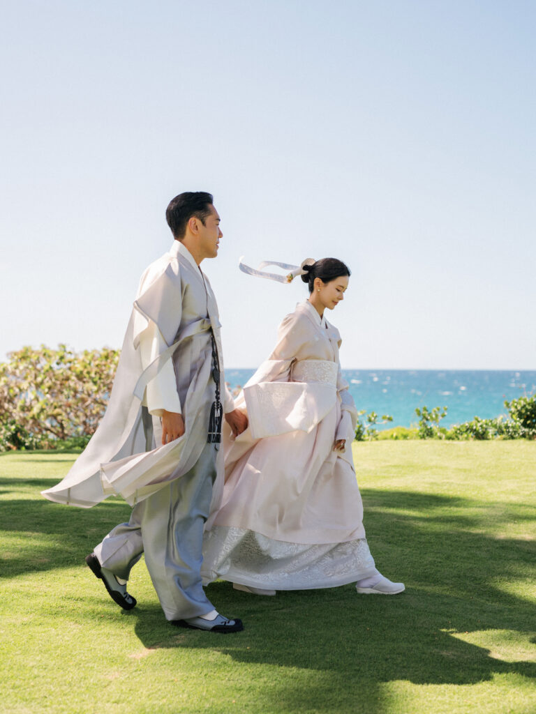 Luxury tropical beach estate wedding look with newlyweds posing in high-end traditional Korean wedding clothes on a green lawn.