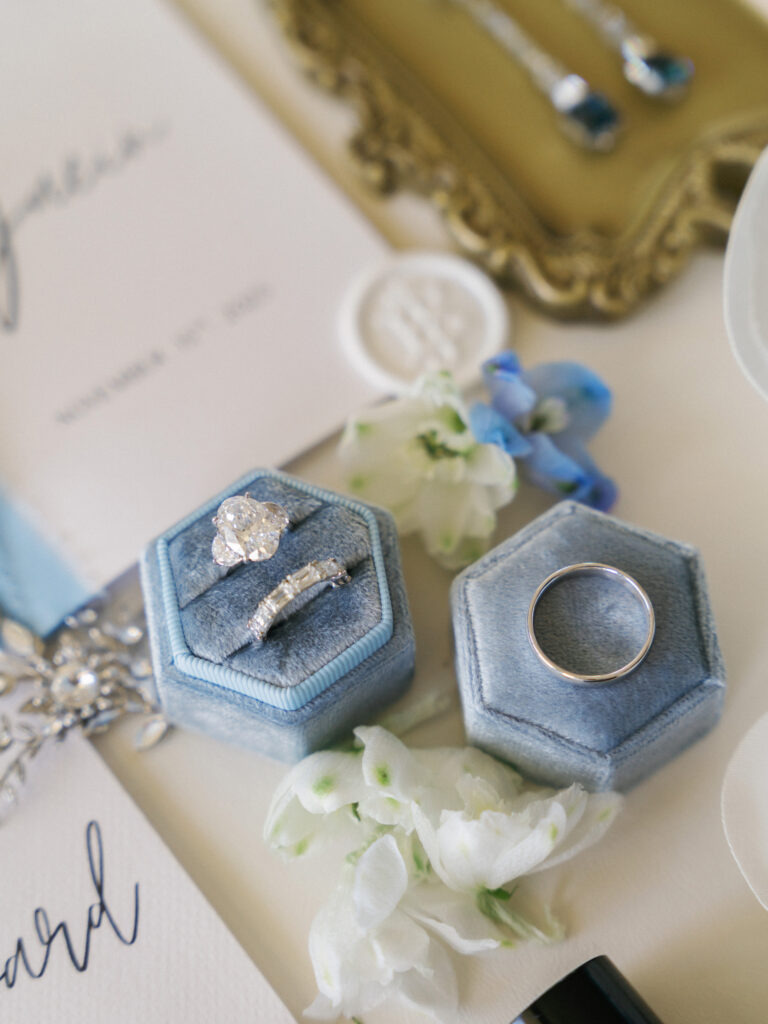 Flat lay of white gold wedding rings in hexagonal velvet boxes, floral accents, and calligraphy invitation cards.