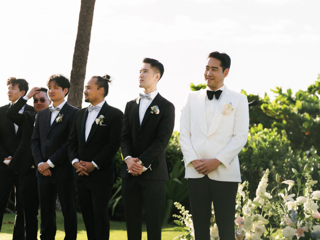 A groom in a white tuxedo jacket and black bow tie standing at the altar, smiling as he watches his bride walk down the aisle during the wedding ceremony.