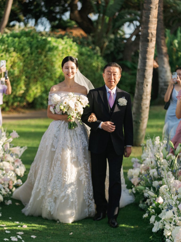 A smiling bride in an off-the-shoulder lace gown holding a bouquet while walking down the aisle with her father during an outdoor wedding ceremony.