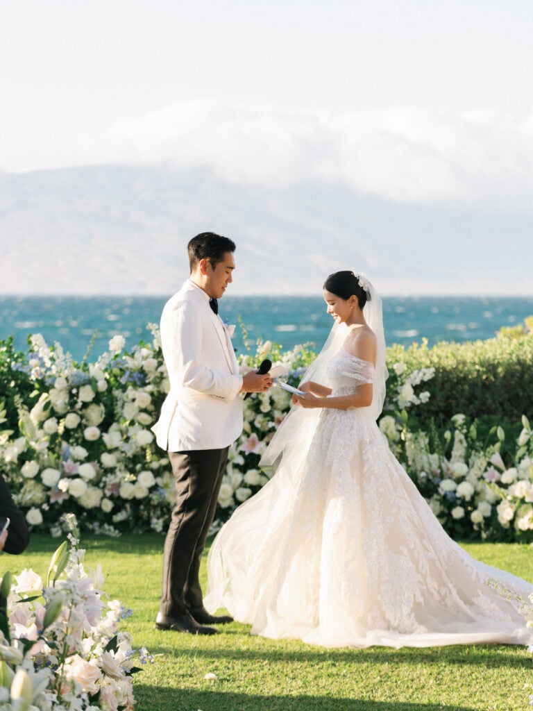 Bride and groom holding hands and standing facing each other at an outdoor wedding ceremony surrounded by white flowers with an ocean background.