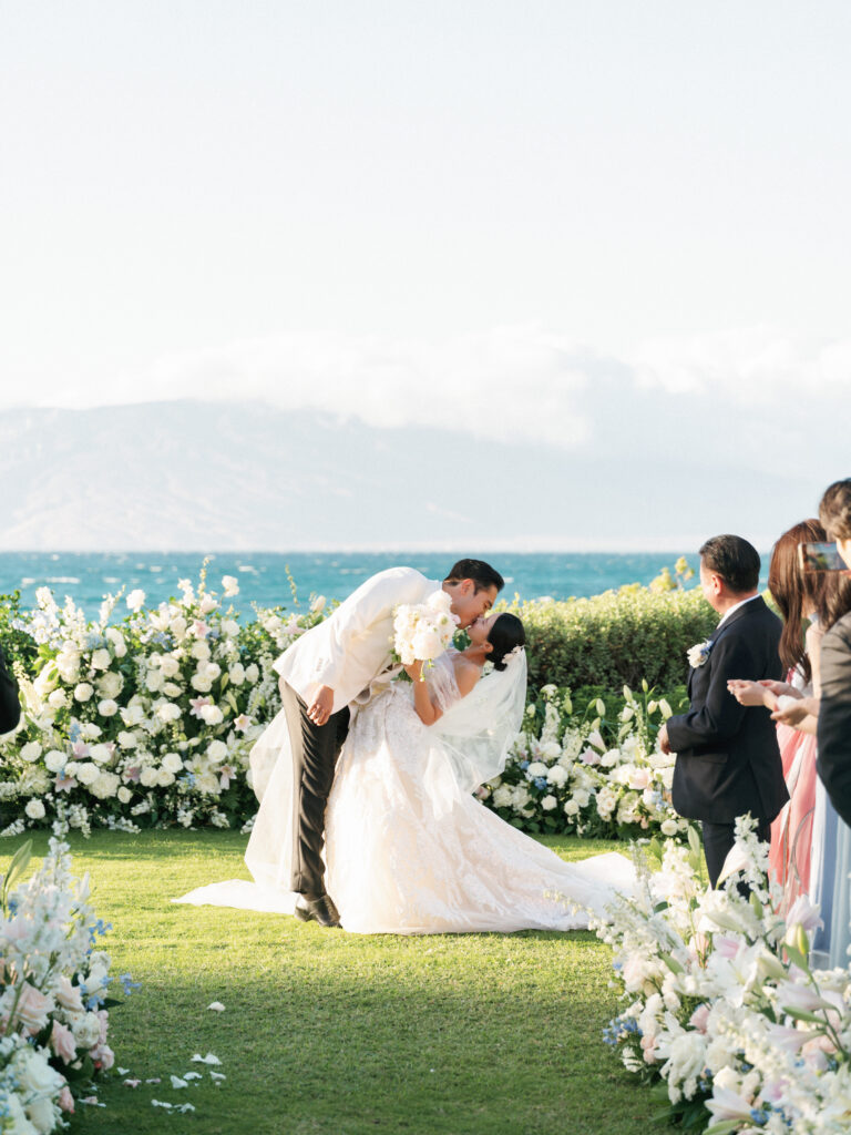 Vintage aesthetic tropical destination wedding featuring a bride in an off-the-shoulder lace dress dipping for a kiss with her groom in a white tuxedo.
