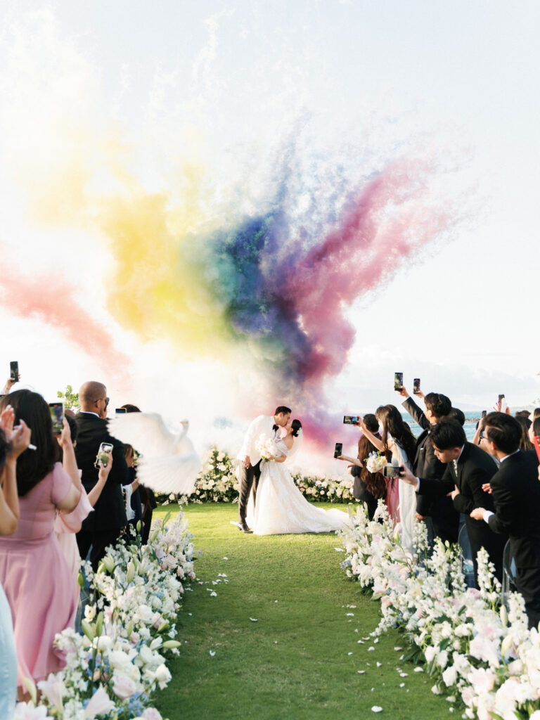 Vintage film aesthetic oceanfront wedding featuring a dramatic rainbow smoke explosion behind the newlyweds kissing at a tropical villa.