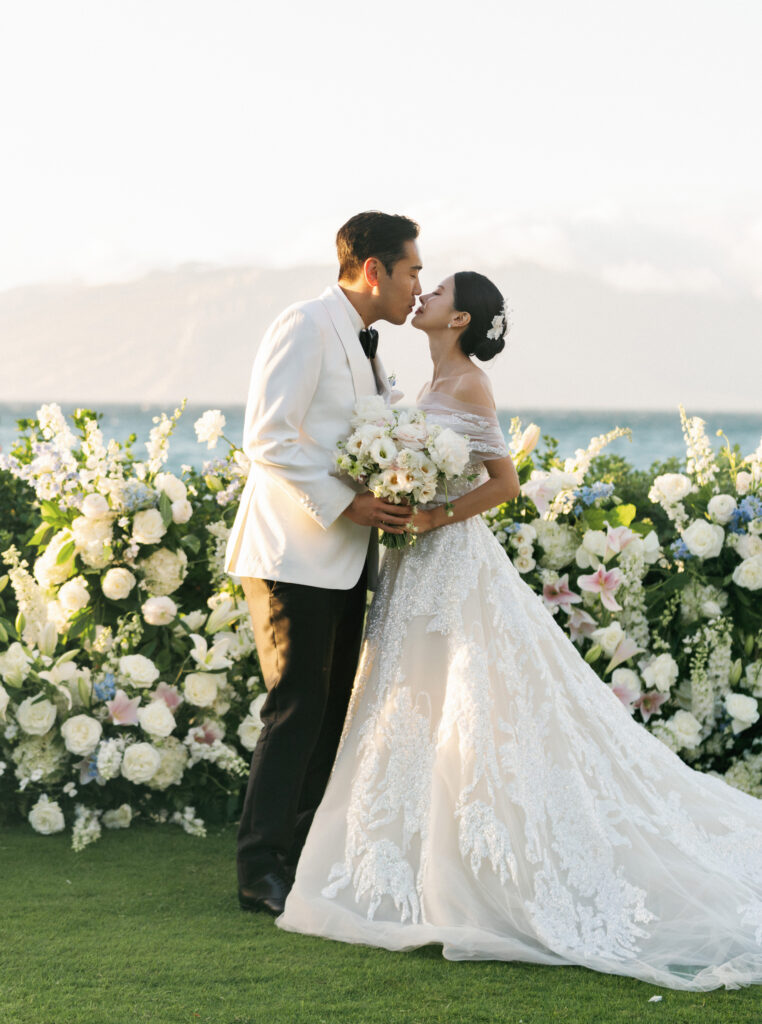 Vintage film aesthetic beachfront wedding ceremony featuring a bride in a lace off-the-shoulder gown and groom in a white tuxedo jacket.