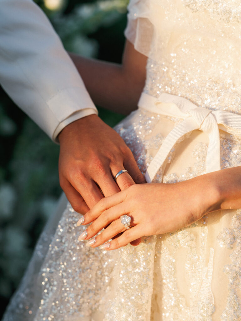 Vintage aesthetic wedding photography detail of bride and groom hands with a luxury oval cut diamond ring against an embellished couture bridal gown.