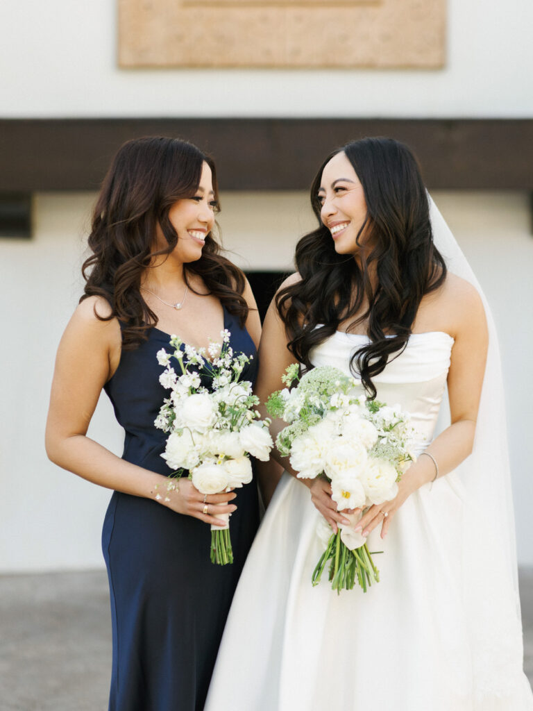 Close-up of a bride and her maid of honor holding organic lush bouquets featuring white peonies, ranunculus, and garden roses.