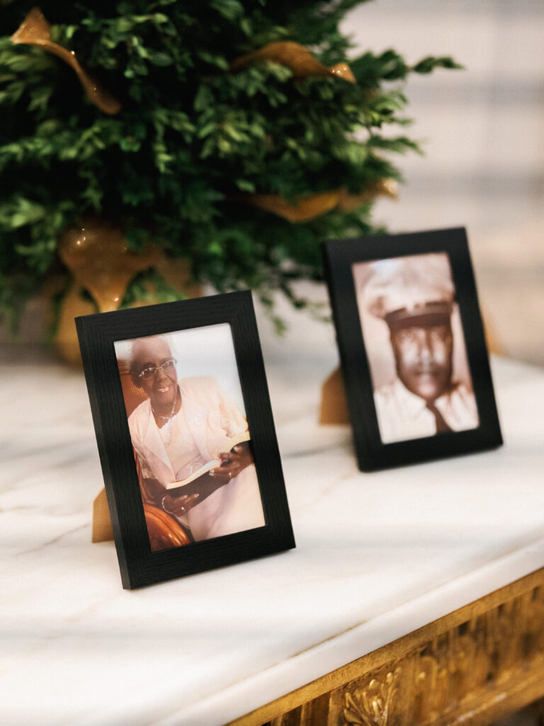 Minimalist wedding memorial display featuring black photo frames of late grandfathers on a luxury white marble table with gold trim next to green topiaries.