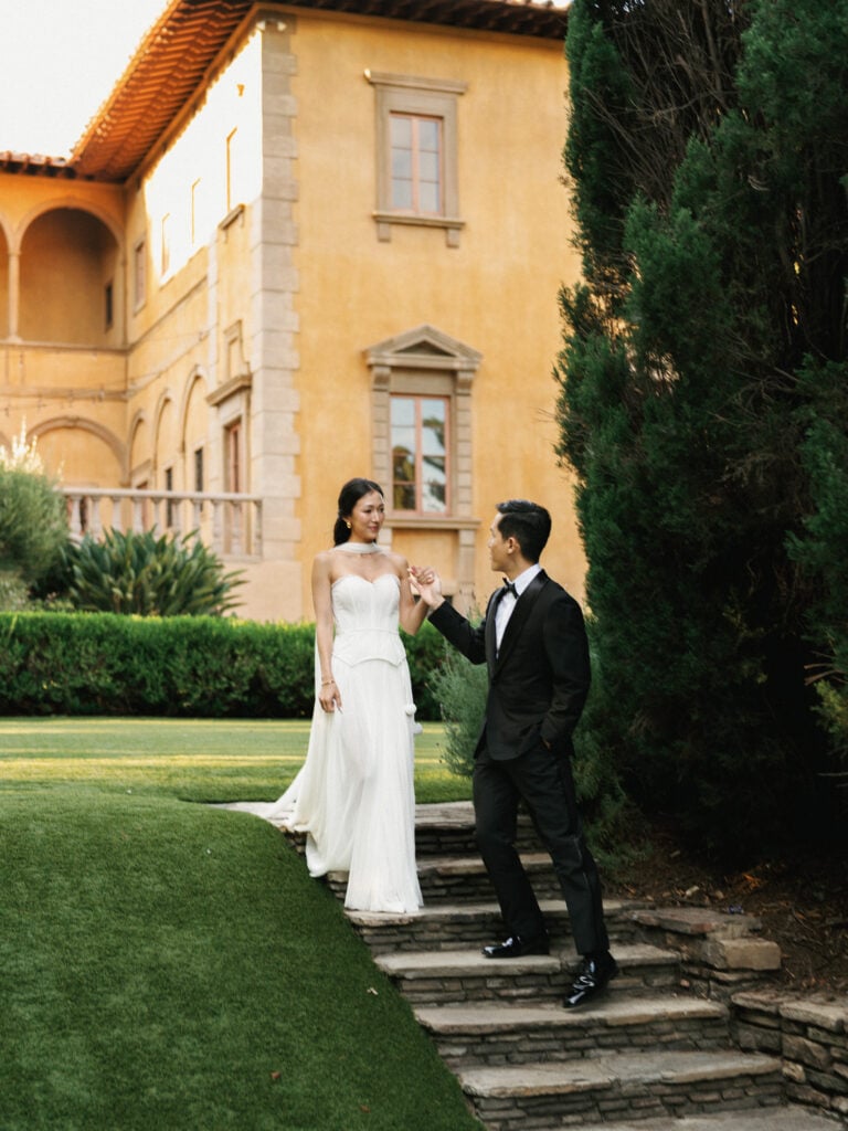 Full-length portrait of a bride and groom walking down stone steps while looking at each other, with a large European-style villa and tall green trees in the background.