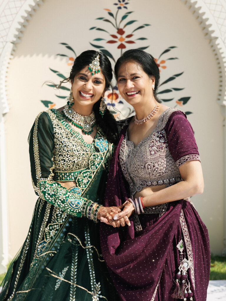 A smiling bride in a green lehenga holding hands with her mother in a purple lehenga at a traditional Indian wedding.