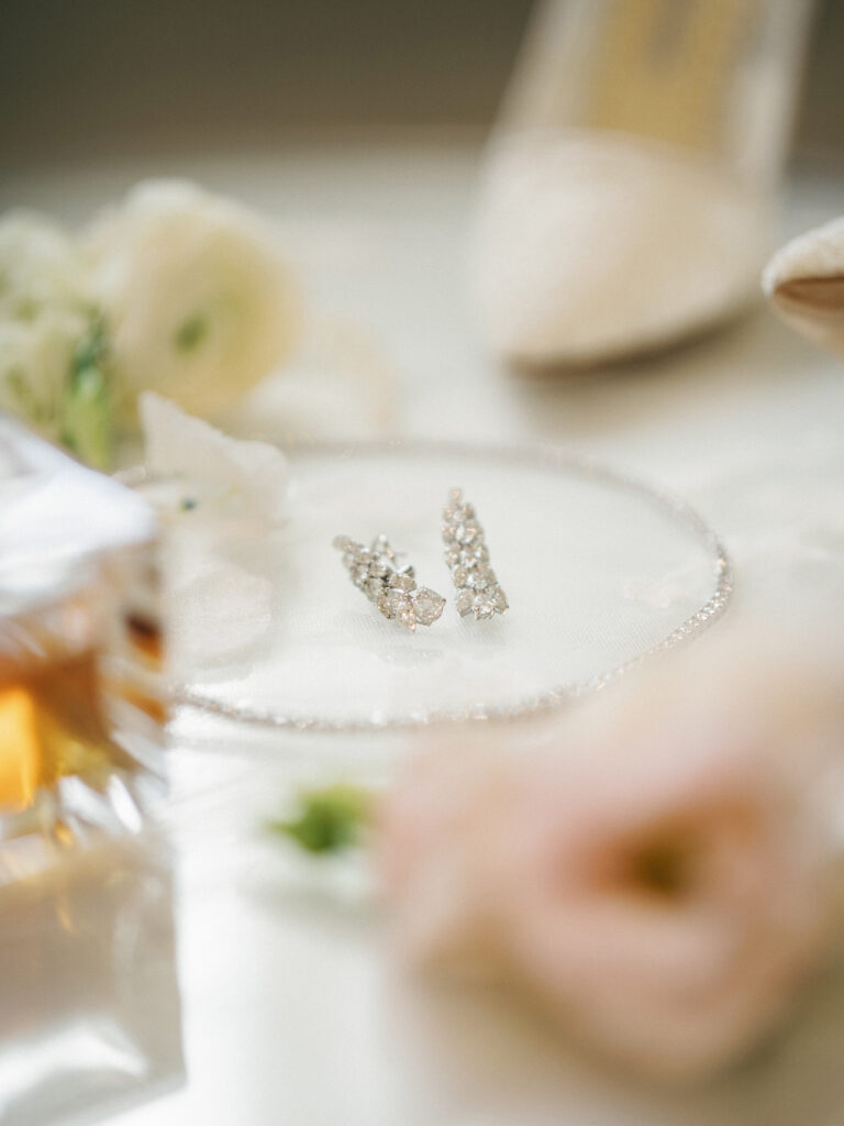 Diamond cluster wedding earrings resting on a small round glass tray next to blurred white ranunculus flowers and bridal heels.