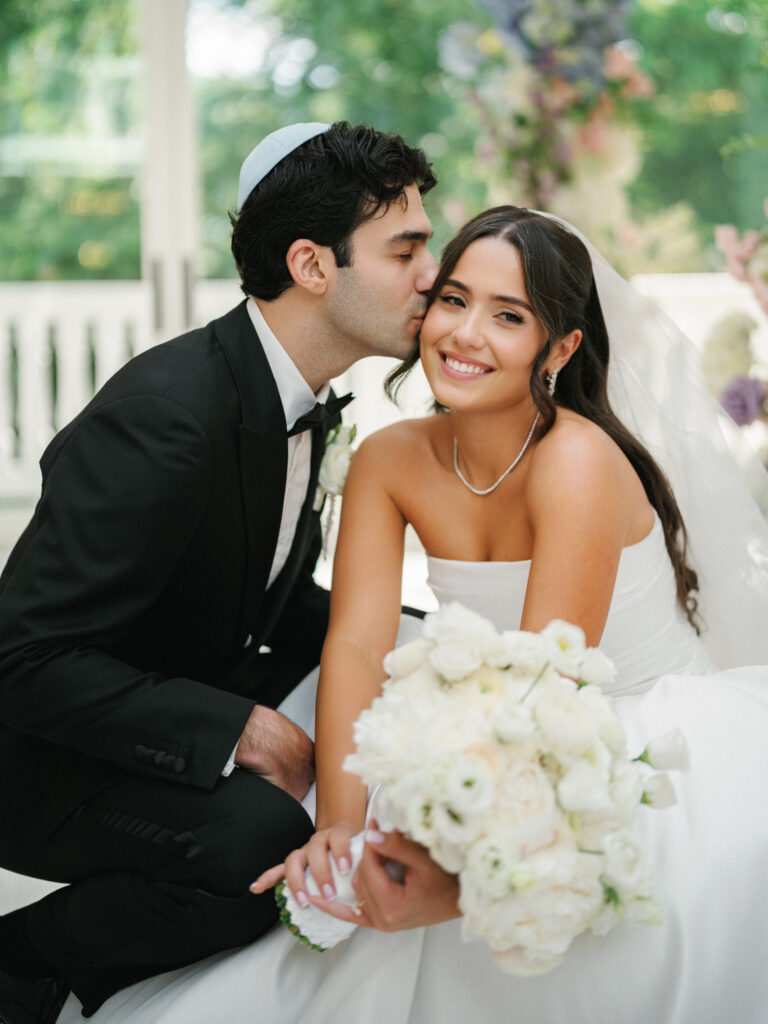 Jewish groom wearing a white yarmulke kissing his smiling bride in a strapless gown, diamond tennis necklace, and long sheer veil.