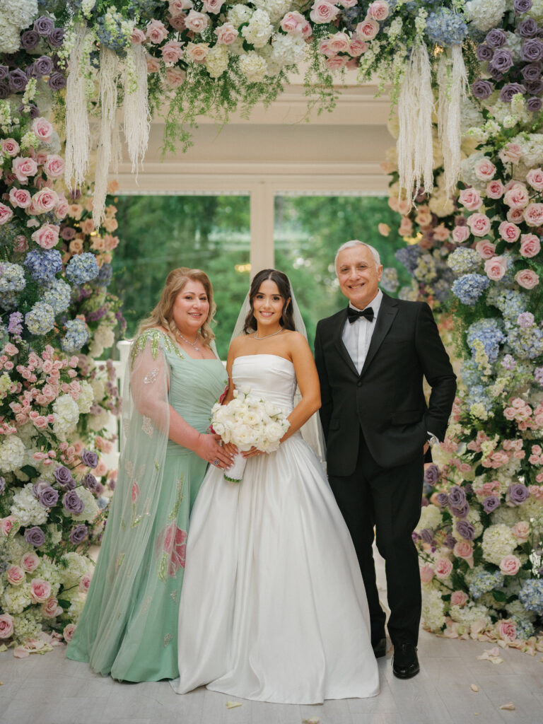 Editorial bride portrait with her parents in front of a lavish indoor flower wall installation filled with pastel roses, hydrangeas, and elegant hanging pampas grass.