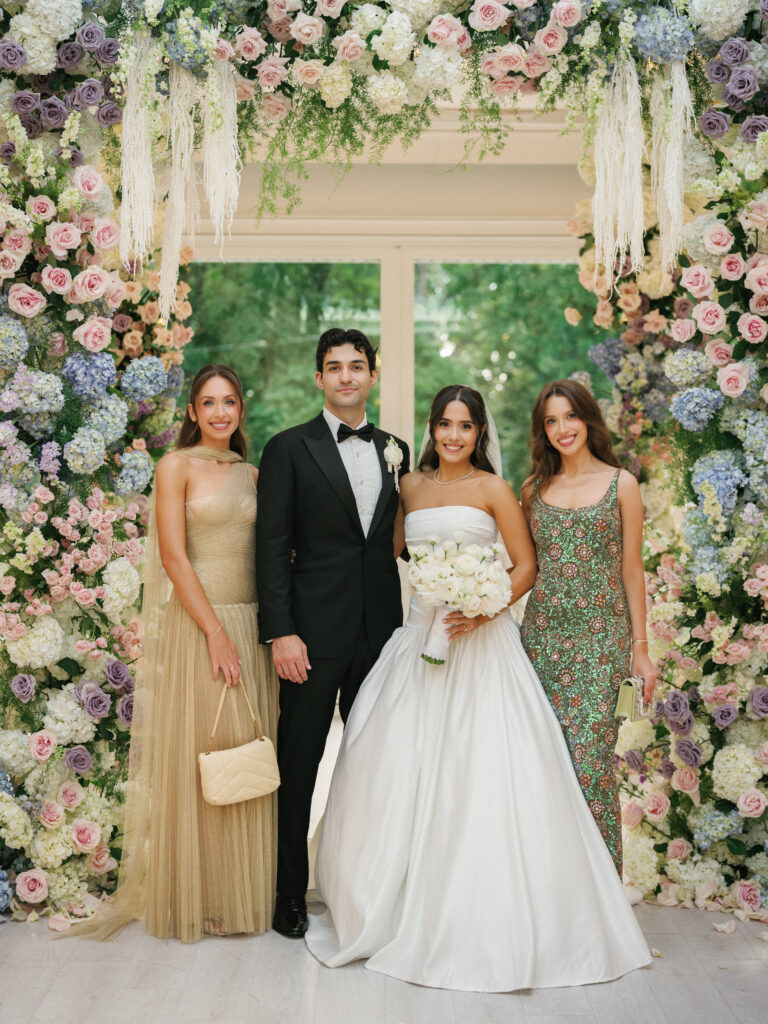 Sophisticated indoor wedding reception photo showcasing newlyweds with friends against a magnificent backdrop of multi-colored pastel floral designs.