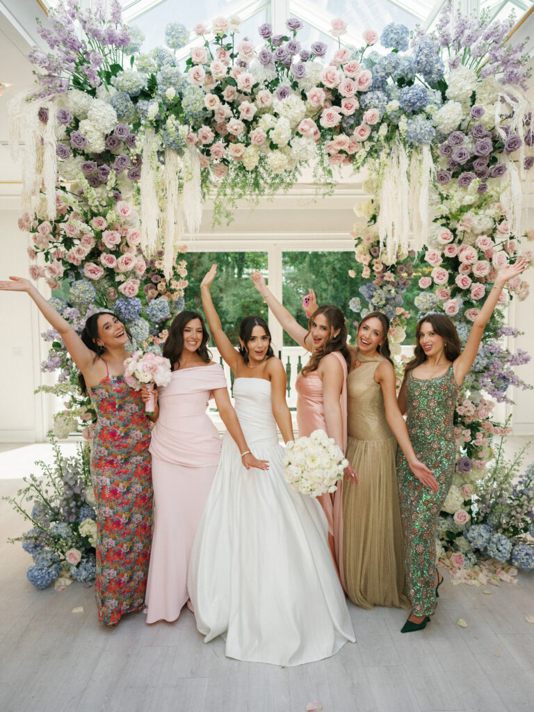 Joyful bride in a strapless white gown celebrating with bridesmaids in mismatched pastel, floral print, and gold dresses in front of a lavish indoor flower arch.
