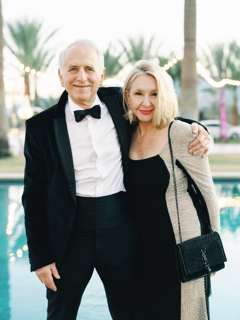 Elegant wedding guests posing by an outdoor pool, featuring a man in a black velvet tuxedo with a bow tie and a woman in a black dress with a YSL chain shoulder bag.