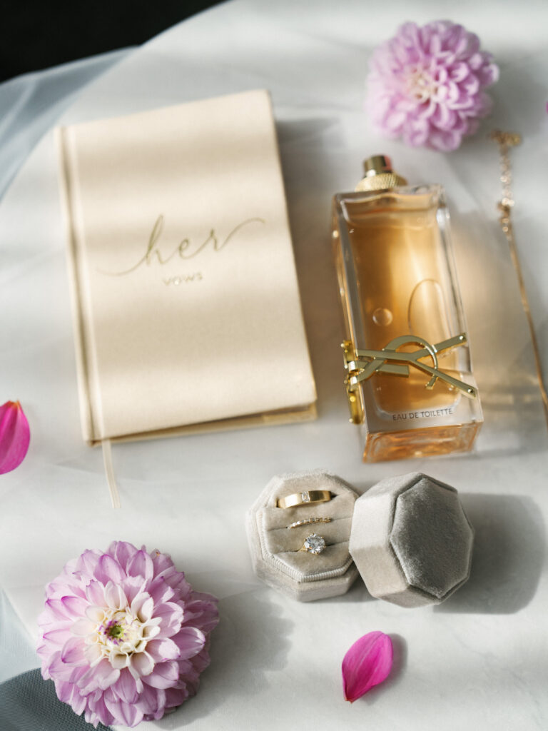 YSL Libre perfume bottle and an ivory velvet Her vows book arranged on a white surface with purple dahlia flowers and a grey hexagon ring box for high-end wedding detail photos.