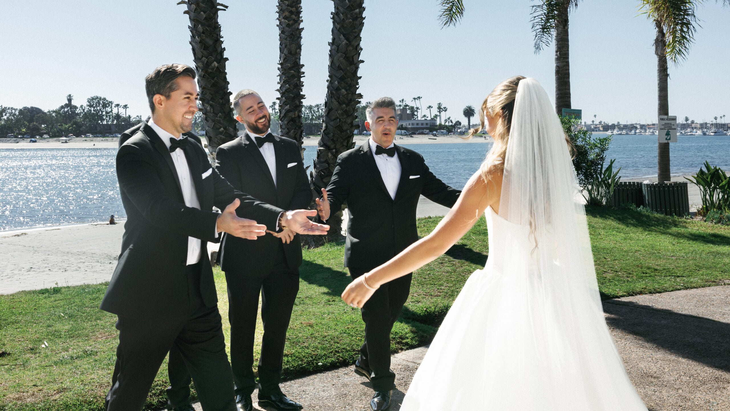 Three groomsmen in black tuxedos gesturing with open arms and smiling in surprise while looking at a bride in a white gown and veil during a pre-ceremony reveal session.