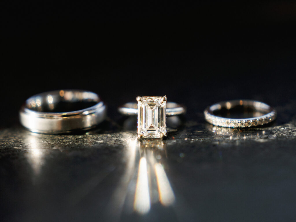 Emerald cut diamond engagement ring with a hidden halo standing on a dark reflective stone surface, casting bright sun flares.