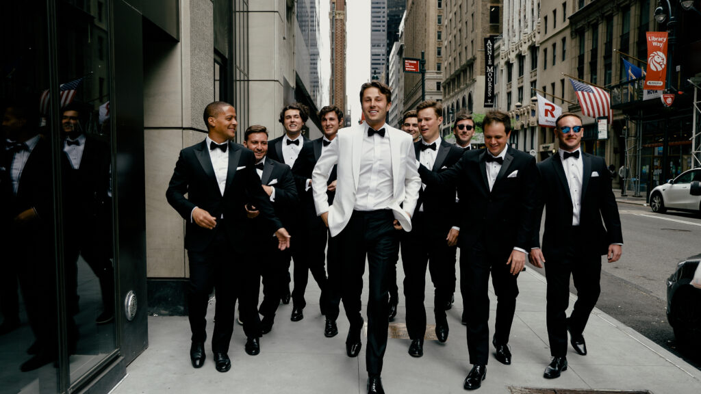 A groom in a white tuxedo walks with his groomsmen in black tie down Library Way in New York City.