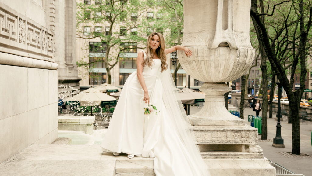 A bridal portrait at the New York Public Library terrace, showcasing a satin ball gown with Bryant Park in the background.