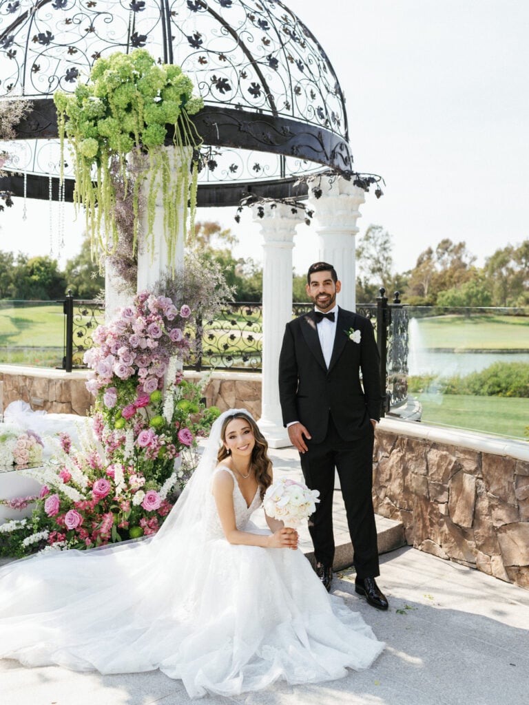 Editorial wedding portrait of a bride and groom posing by a floral gazebo at a multicultural Persian wedding.