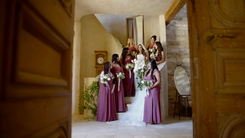 Full-length front view of a bride and bridesmaids in mauve floor-length dresses posing on a curved stone staircase inside Sunstone Villa, holding classic white and green bouquets.