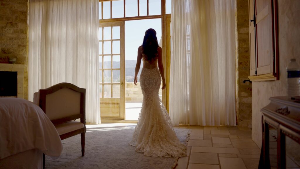 Full-length back view of a bride in a luxury floral lace mermaid wedding dress walking toward open French doors inside a sunlit suite at Sunstone Villa.