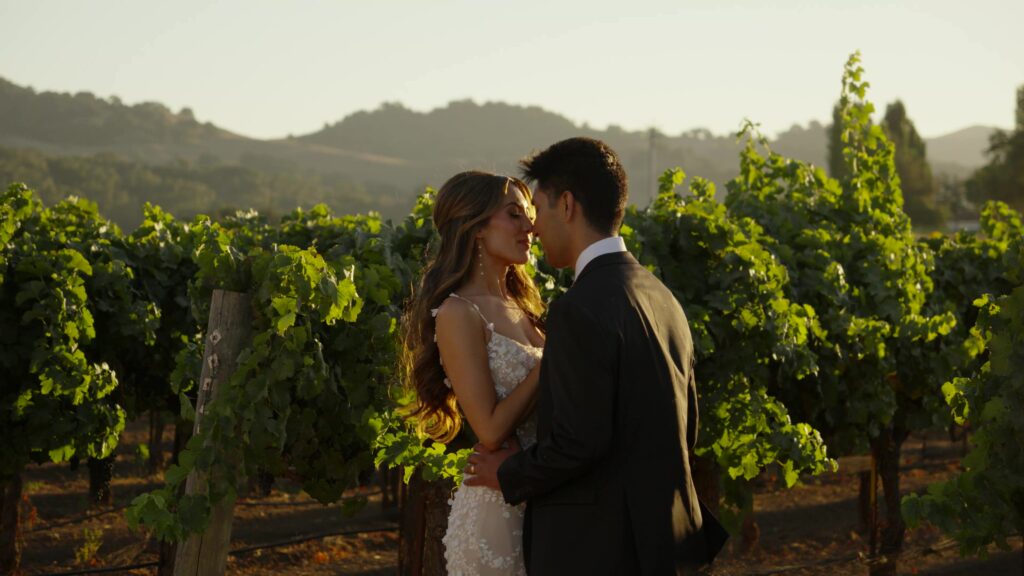 Cinematic golden hour wedding portraits at Sunstone Villa, showcasing an intimate couple embrace standing between rows of lush green grapevines in the Santa Ynez winery.