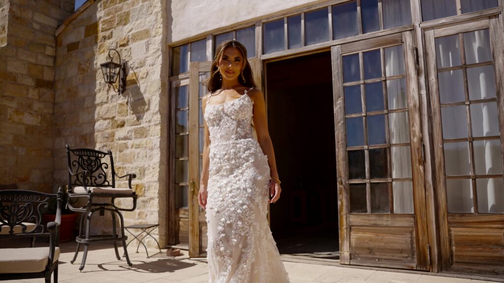 Full-length front view of a bride posing on a stone patio outside Sunstone Villa, wearing a luxury floral applique wedding dress next to rustic wooden French doors.