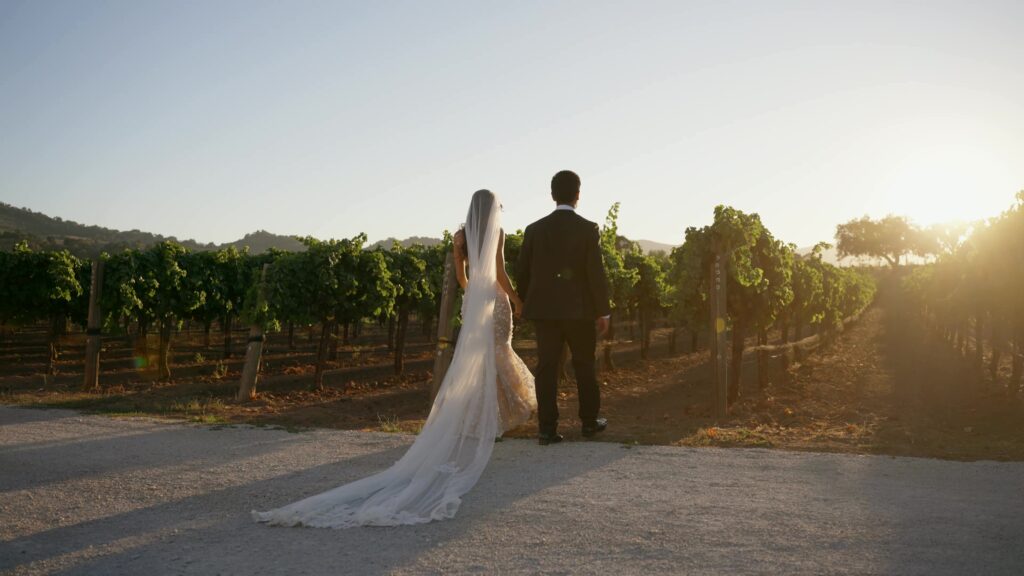 Editorial back view of a bride and groom walking away on a wide gravel road, showcasing a long cathedral veil and sleek gown trailing behind as they face the sunset vineyards at Sunstone Villa.
