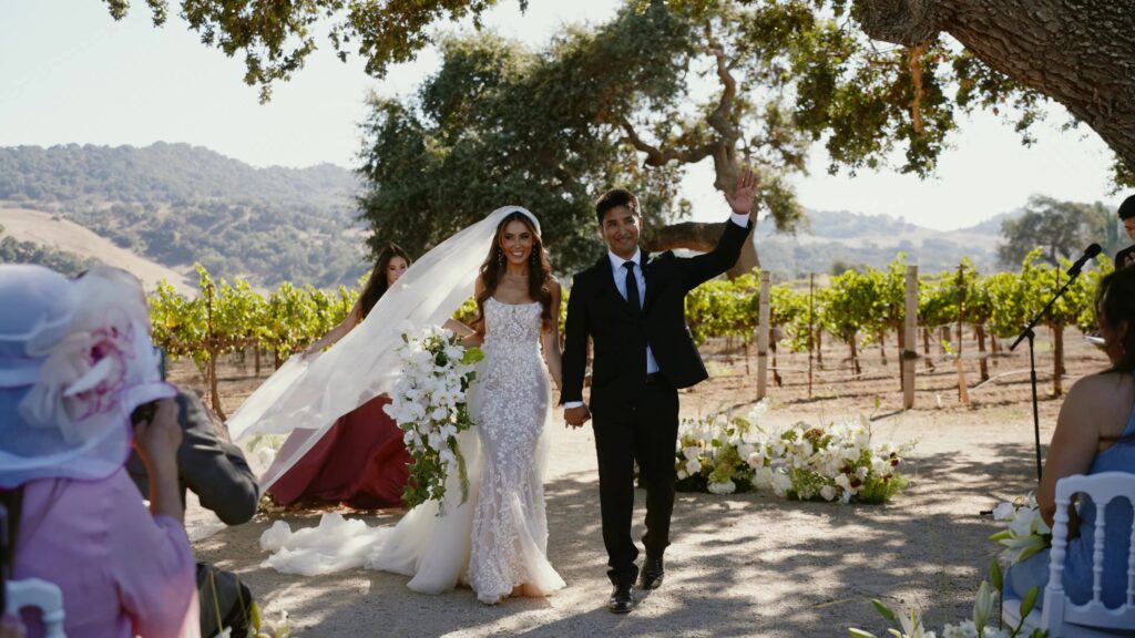 Bride and groom smiling and waving during their wedding recessional at Sunstone Villa, showcasing a white orchid cascading bouquet and lace mermaid dress with Santa Ynez vineyards in the background.