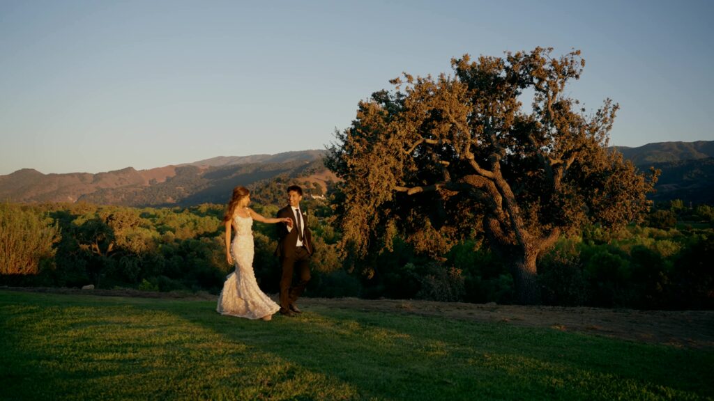 Side profile of a bride and groom holding hands and walking across a manicured green lawn at Sunstone Winery, with a large historic oak tree and rolling hills in the background during golden hour.
