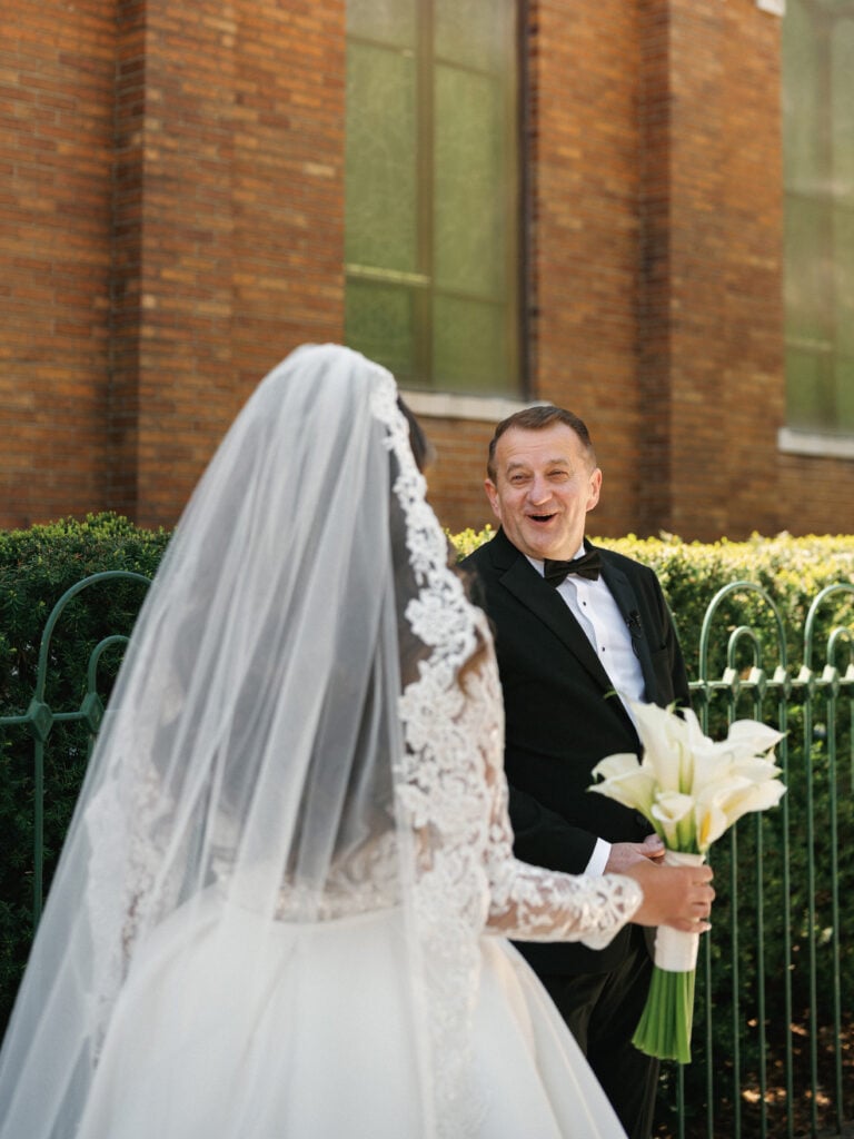 An emotional father in a black tuxedo smiling with surprise while looking at his bride daughter during a father first look reveal session outdoors.