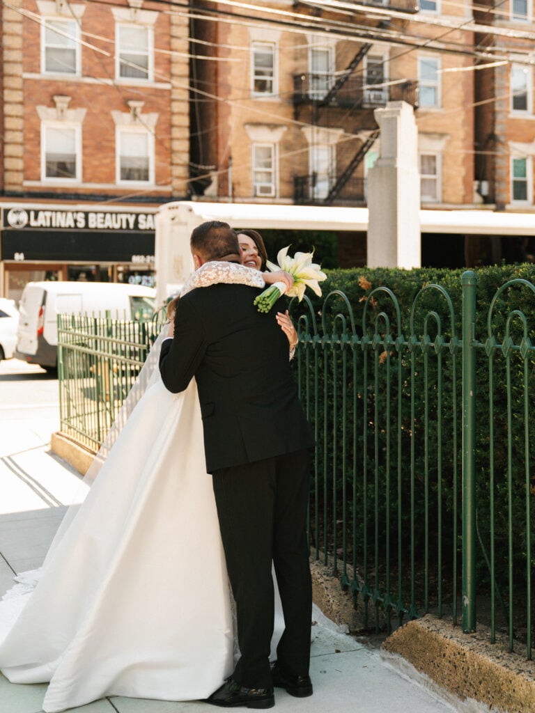 A bride in a white wedding gown holding a calla lily bouquet hugs her father in a black tuxedo during an emotional father first look reveal on a city sidewalk.